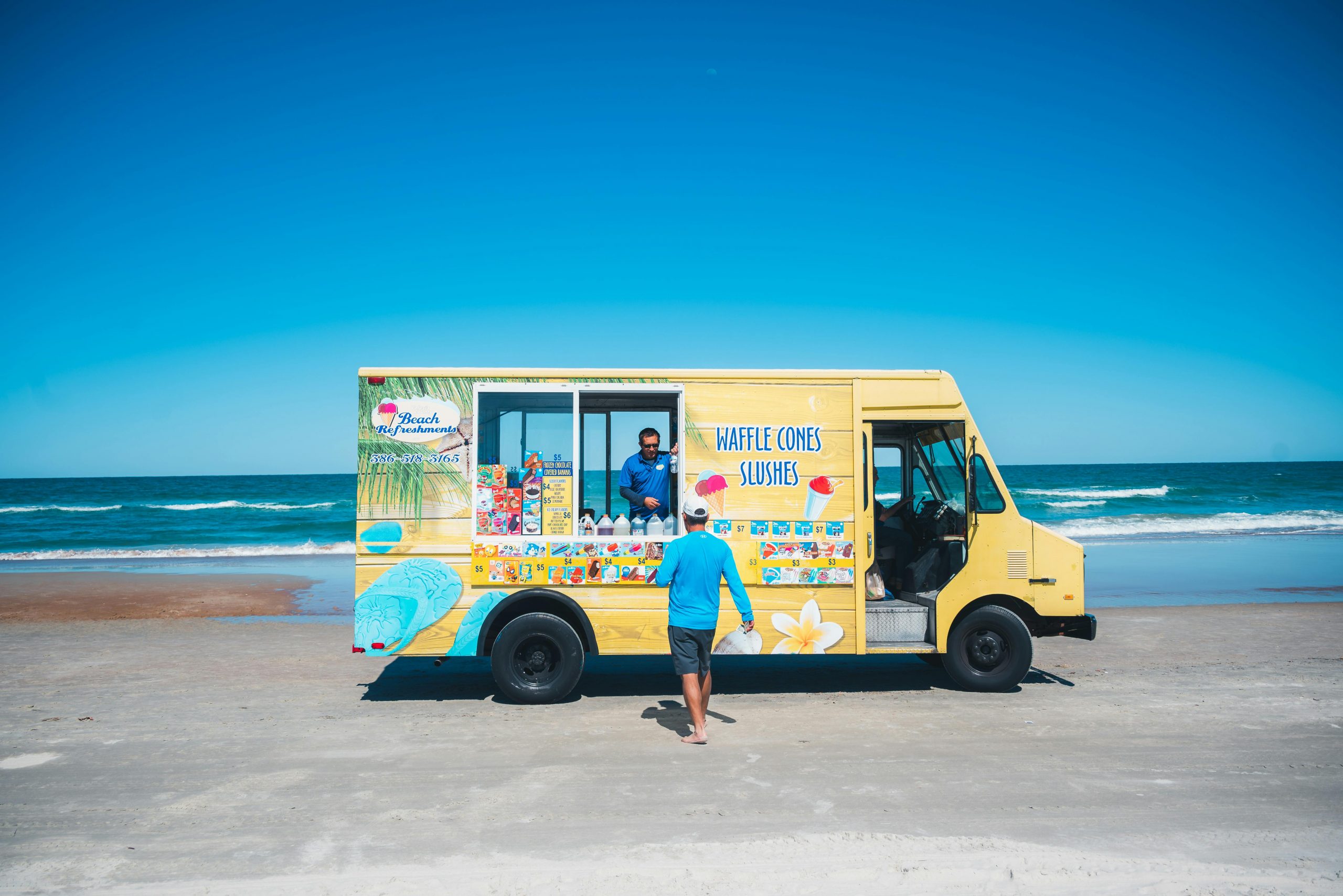 A bright yellow ice cream truck on a sunny beach, serving treats under a clear blue sky.