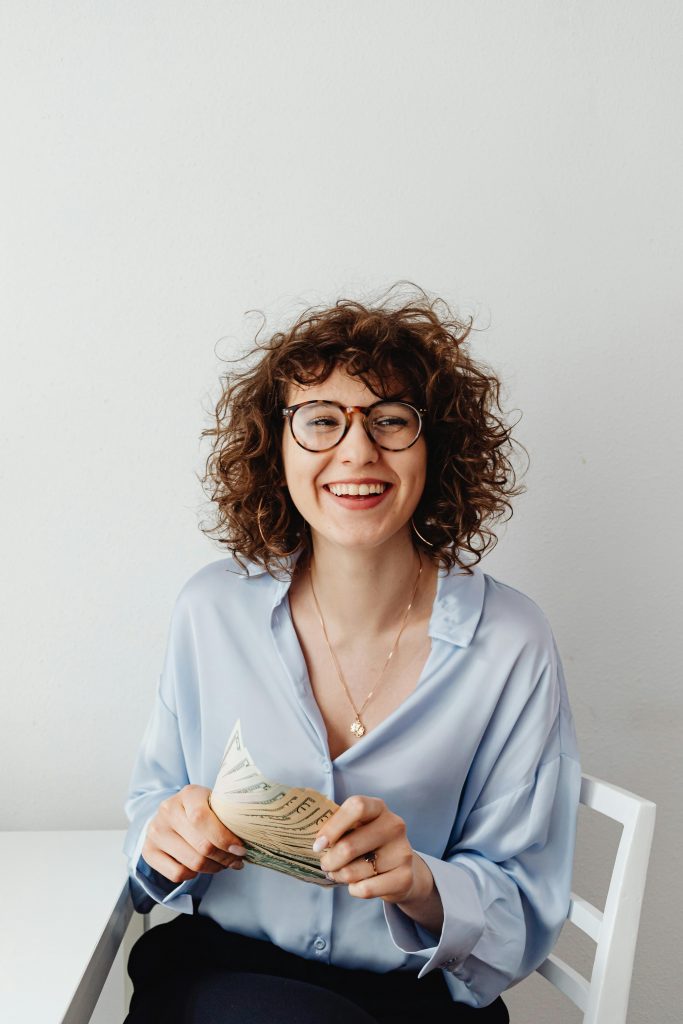 pexels-photo-7680629-7680629 Cheerful woman with curly hair and glasses holding US dollars, sitting indoors.