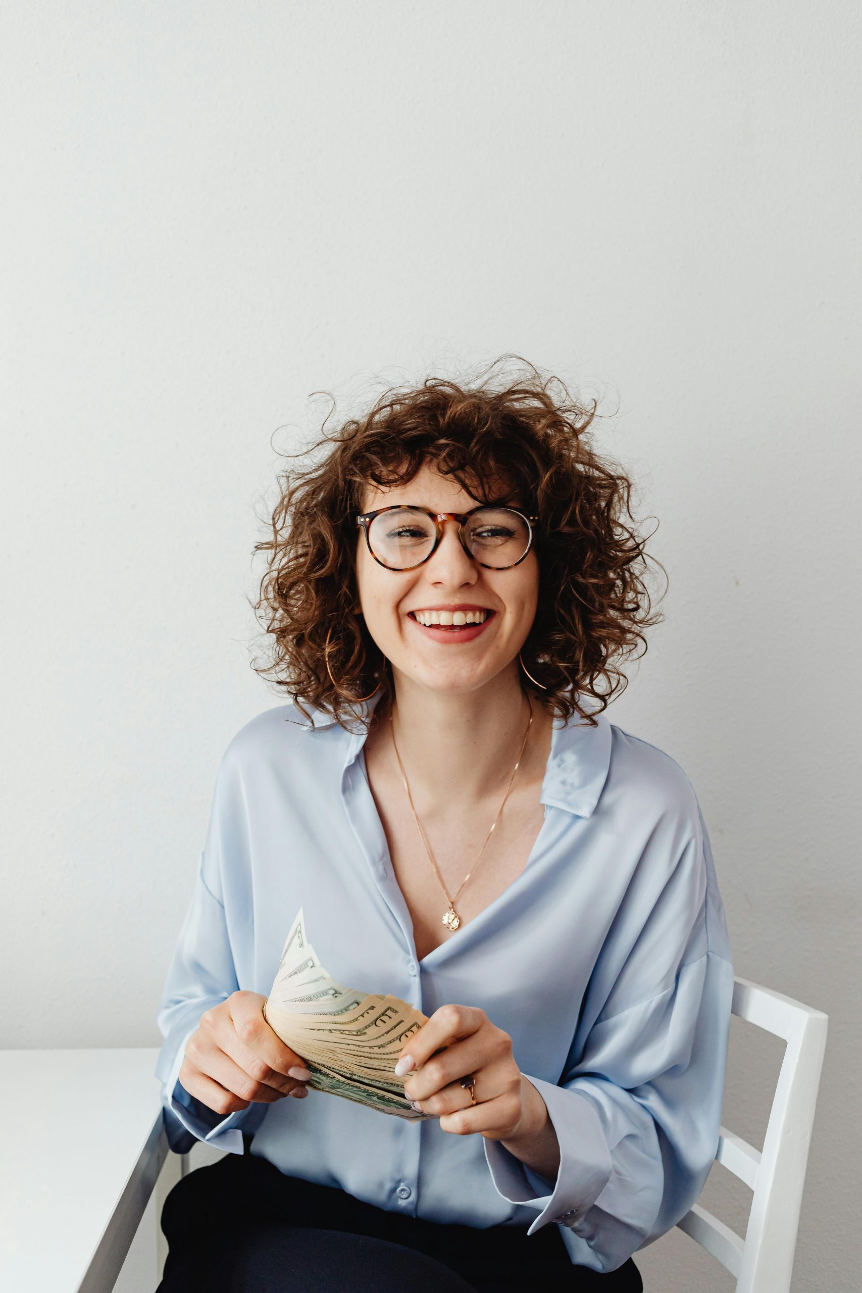 Cheerful woman with curly hair and glasses holding US dollars, sitting indoors.