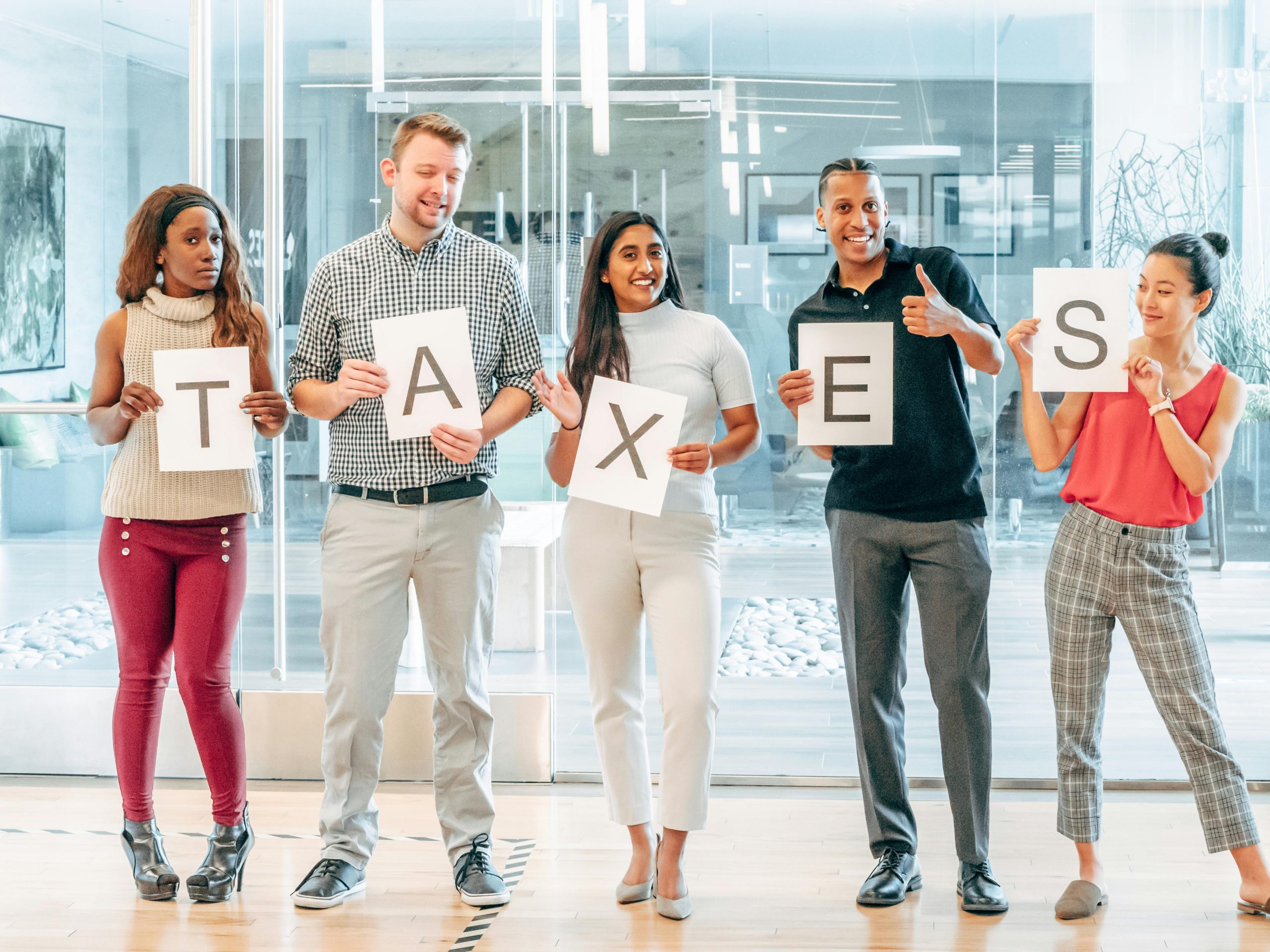 A diverse group of adults holding letters spelling 'taxes' in an office setting.