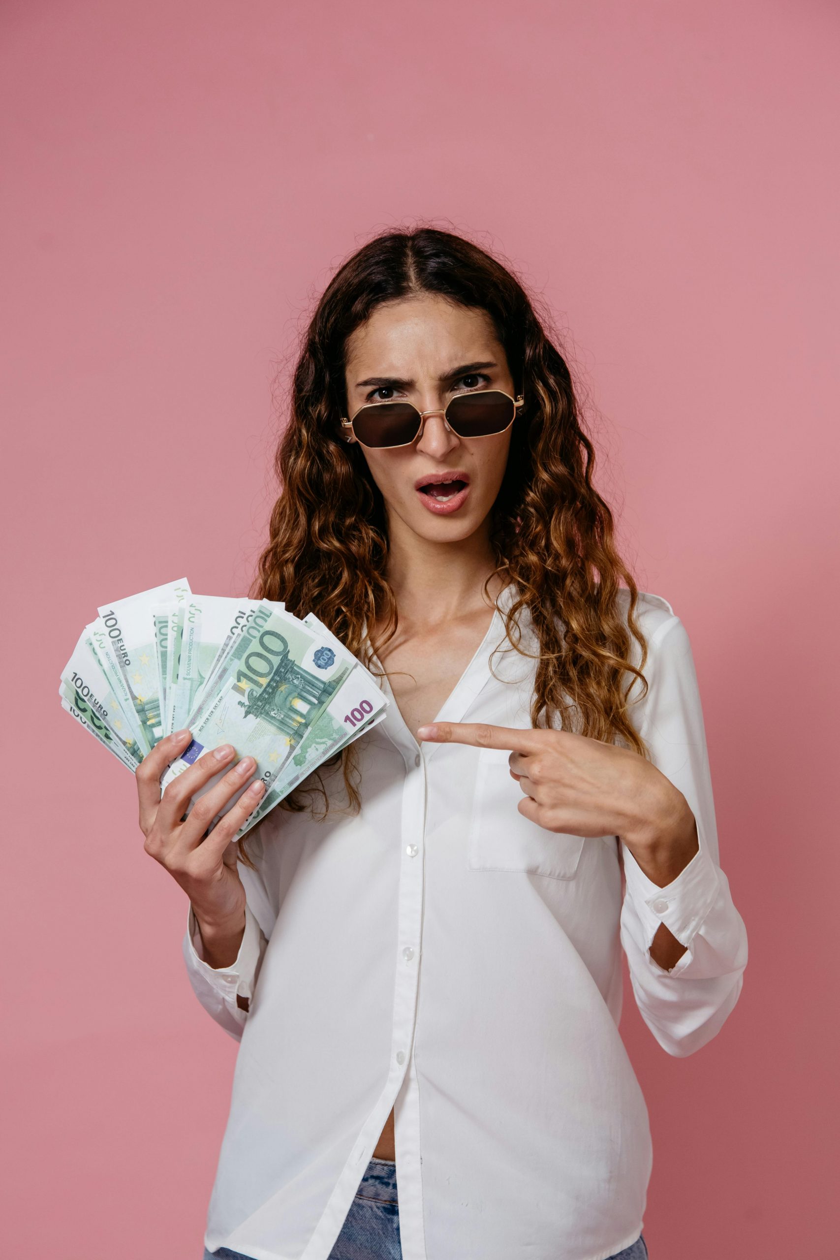 Young woman in sunglasses holding euro banknotes and pointing, pink background