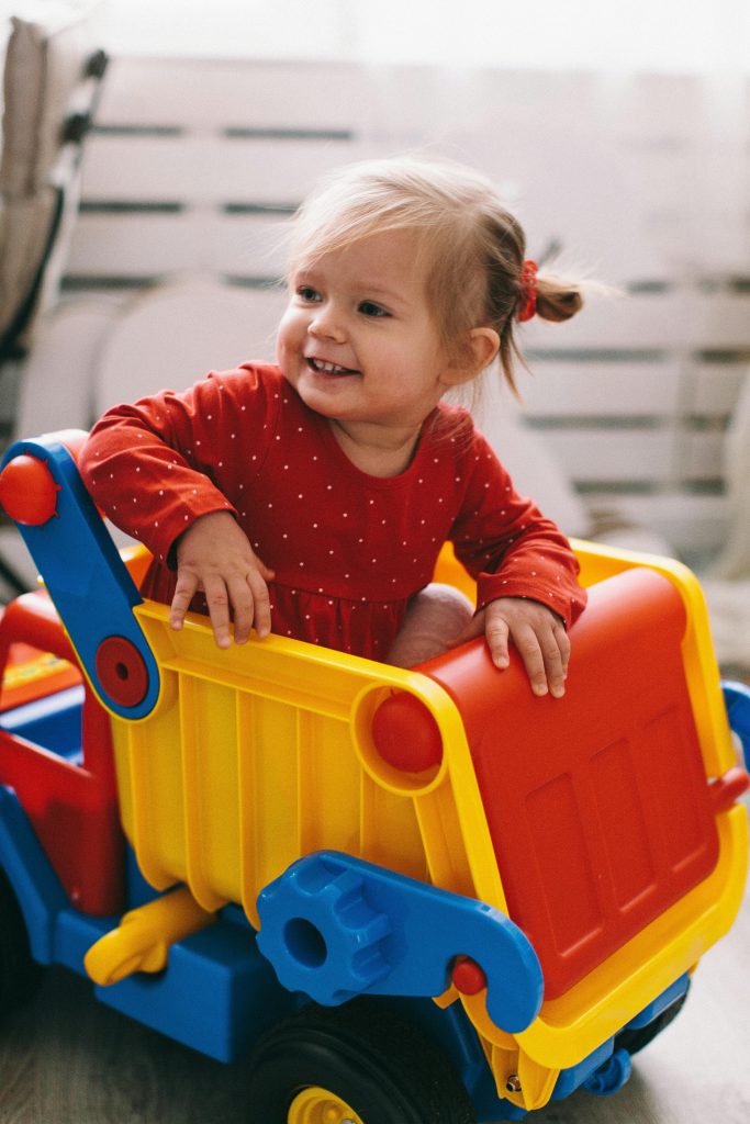 Delighted toddler in a red dress playing with a colorful toy truck indoors.