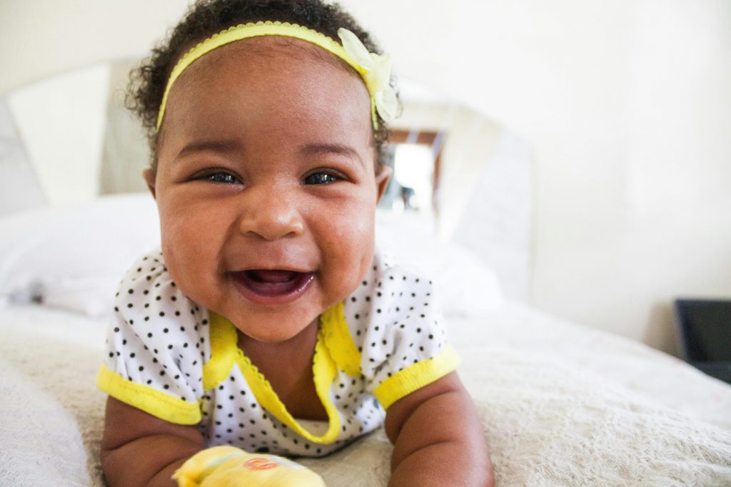 Cheerful baby girl in a polka dot outfit with a big smile on a bed indoors.