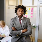 Businessman with dreadlocks in gray suit smiling confidently at office desk.